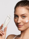 Exuviance kvinna ler mot kameran med kräm på kinden och produkten daily corrector SPF 35 i handen.