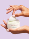 Exuviance produkten skinrise morning glow är placerad i en hand och en annan plockar upp en bomullsrondell från förpackningen.
